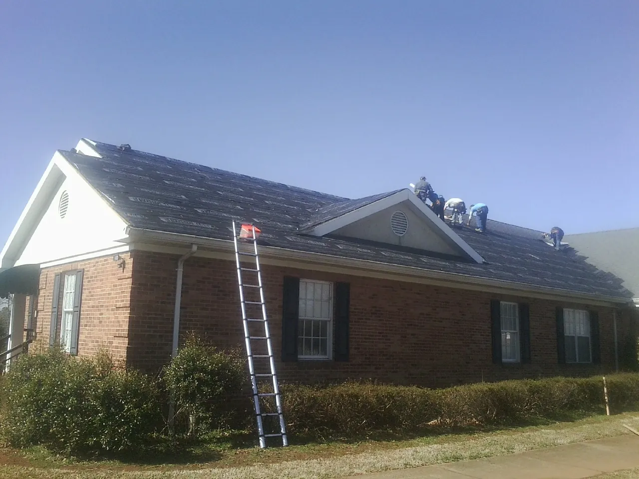 Crew tearing shingles off a brick ranch roof with a ladder set — Emergency Roof Repair work by our crew in Hartington