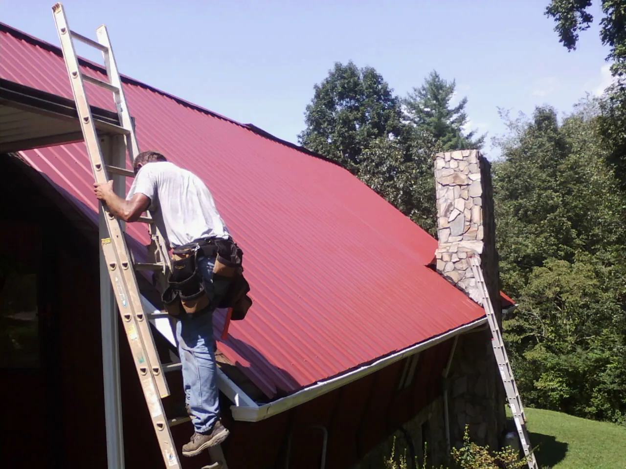 Roofer on a ladder beside a red metal roof and stone chimney — Soffit & Fascia Repair by Roofing Brothers in Hartington